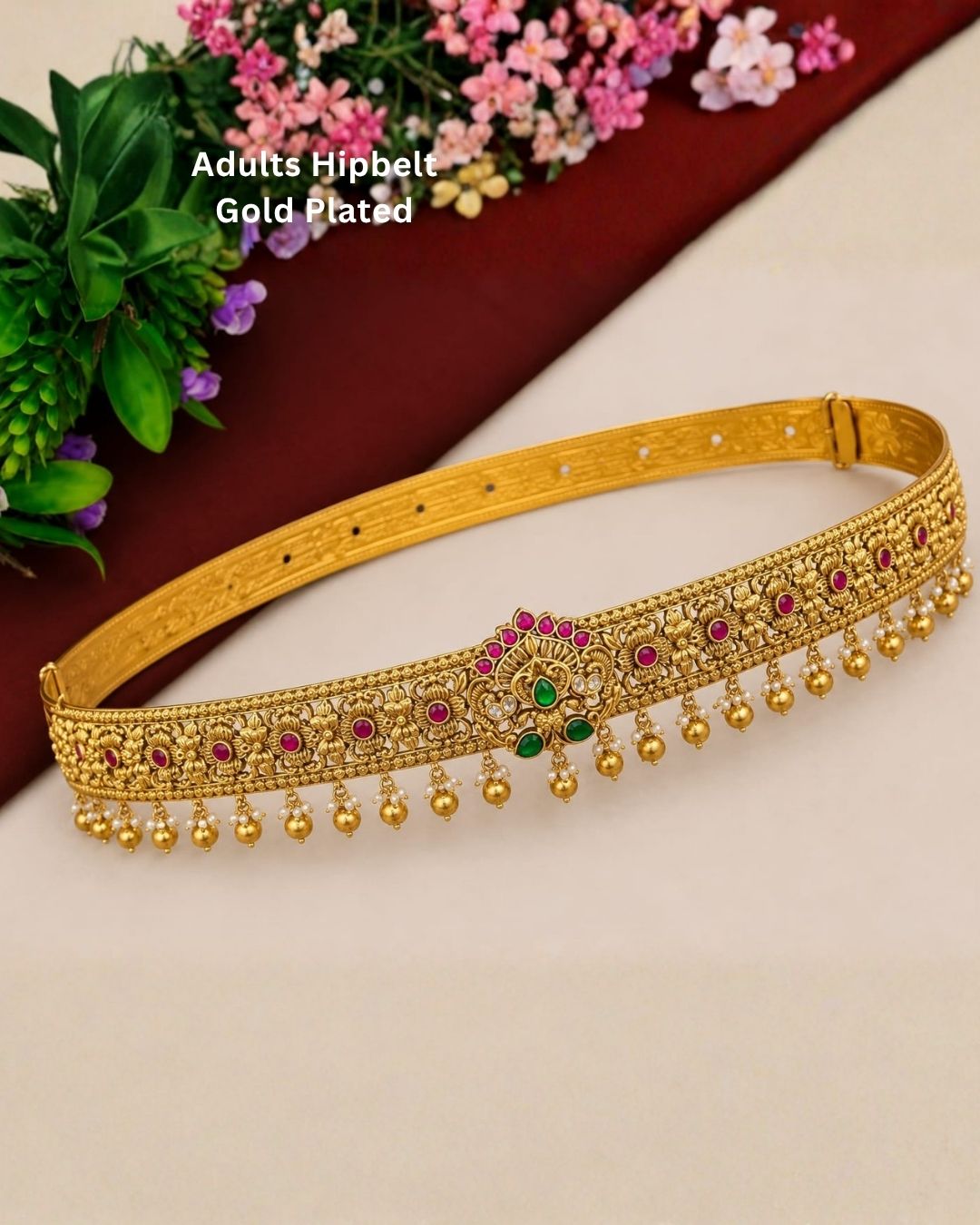 Premium gold plated designer hip belt featuring detailed floral motifs, ruby-colored stone accents, green stones at the center, and elegant hanging gold beads. Perfect for weddings, festive occasions, traditional wear, bridal styling, parties, and gifting.