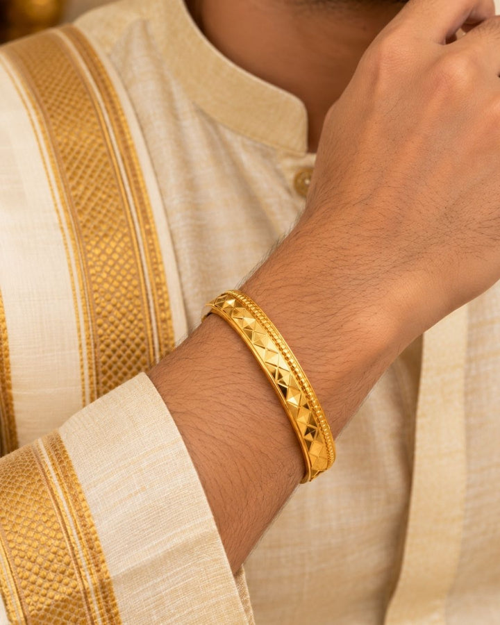 Gold plated men’s kada displayed upright on a warm beige surface, showcasing its diamond-cut design and glossy finish.