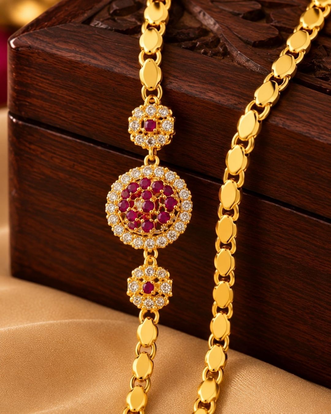 Designer gold plated thalli chain displayed on beige silk with a carved wooden jewellery box, featuring a round ruby stone centre design and matching ornamental accents.