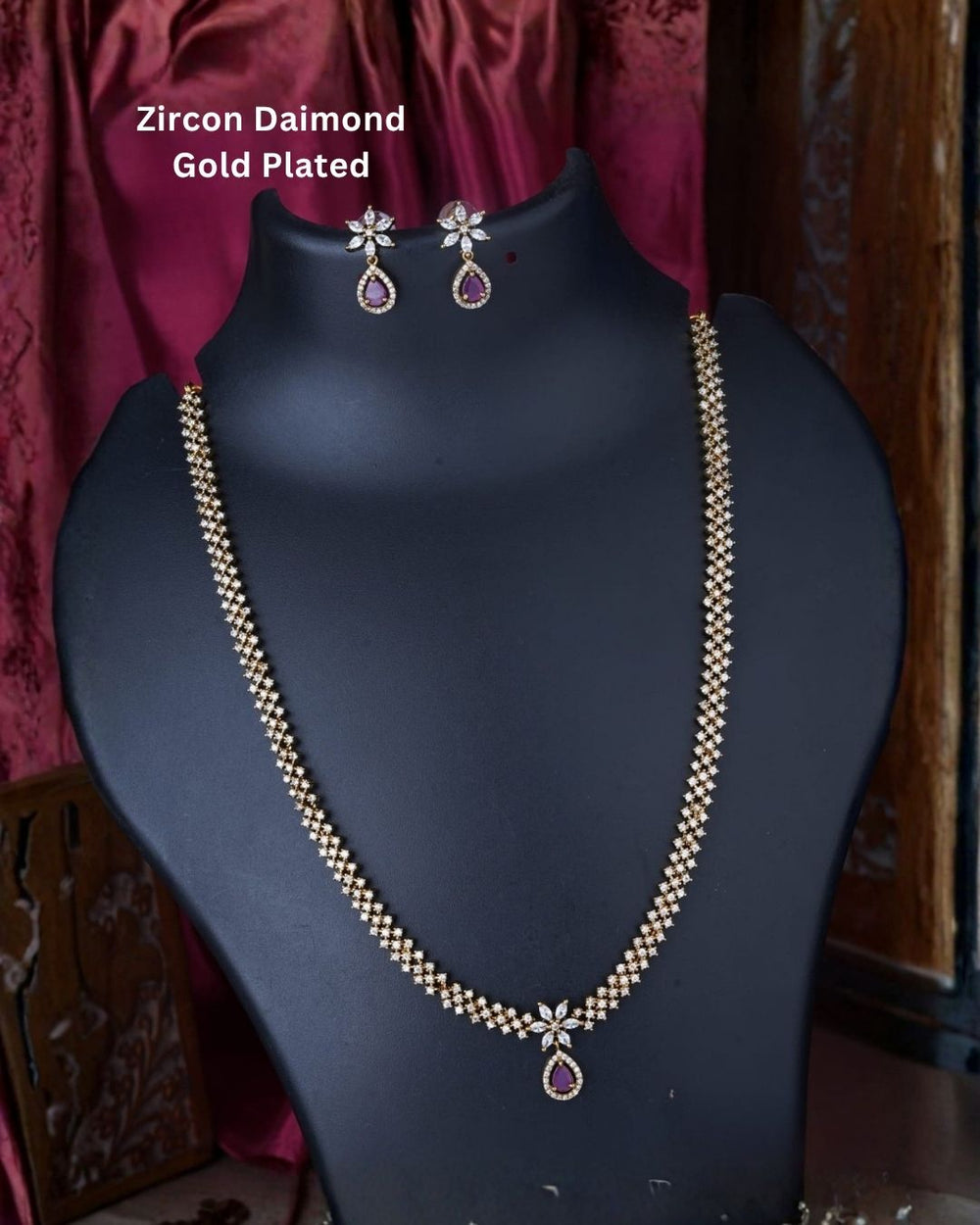 Gold plated zircon necklace and matching earrings displayed on a black jewellery bust against a rich burgundy background.