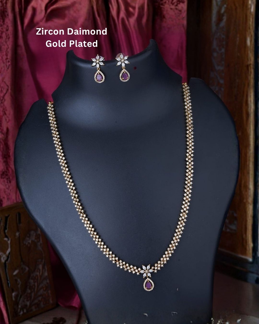 Gold plated zircon necklace and matching earrings displayed on a black jewellery bust against a rich burgundy background.