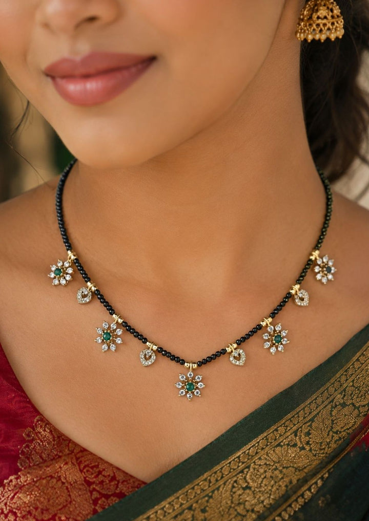 A close-up photo of a south indian woman wearing a beautiful short black beads chain, also known as a mangalsutra. The necklace is made of small black beads with tiny gold dividers. It has pretty charms hanging from it. Some charms look like little flowers with white shiny stones and a green stone in the middle. Other small charms look like shiny little hearts.