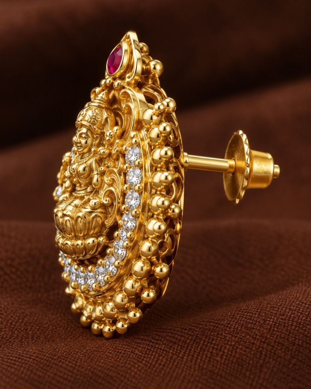 Close-up of South indian women ear wearing a Traditional Laxmi gold plated stud earrings . The circular ear studs feature a highly detailed embossed figure of Goddess Lakshmi sitting on a lotus in the center. The idol is semi-circled by shimmering white CZ American diamond stones, a beaded gold border, and a small red ruby stone at the very top tip. This temple jewelry piece provides an elegant ethnic look suitable for puja festivals, weddings, and traditional family occasions. 