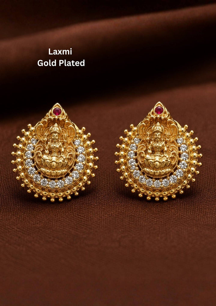 Traditional Laxmi gold plated stud earrings . The circular ear studs feature a highly detailed embossed figure of Goddess Lakshmi sitting on a lotus in the center. The idol is semi-circled by shimmering white CZ American diamond stones, a beaded gold border, and a small red ruby stone at the very top tip. This temple jewelry piece provides an elegant ethnic look suitable for puja festivals, weddings, and traditional family occasions. 