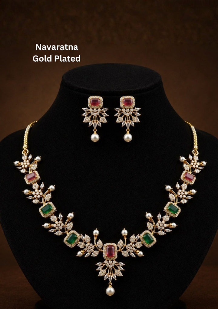 Gold-plated Navratna jewelry set featuring a necklace and matching earrings The set is adorned with gold-plated floral motifs, green rectangular stones, pink rectangular stones, and white pearl-like drops. Elegant Navaratna gold plated necklace set featuring colorful gemstone-inspired stones, pearl drops, and matching earrings for festive occasions.