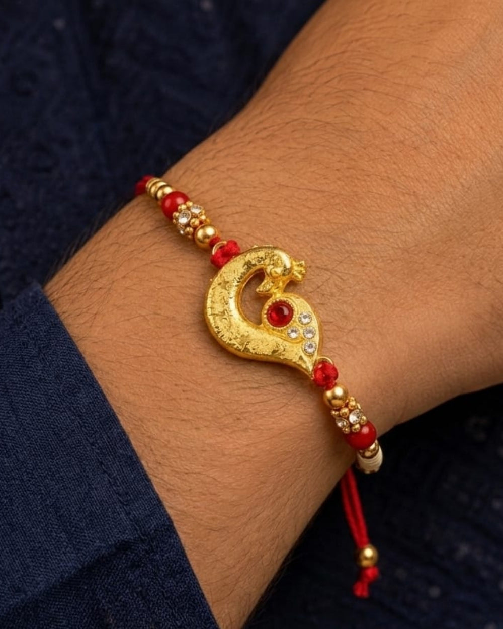 Elegant gold plated Rakhi showcasing a peacock-inspired centerpiece with red stone embellishments, zircon detailing, and a vibrant red thread.