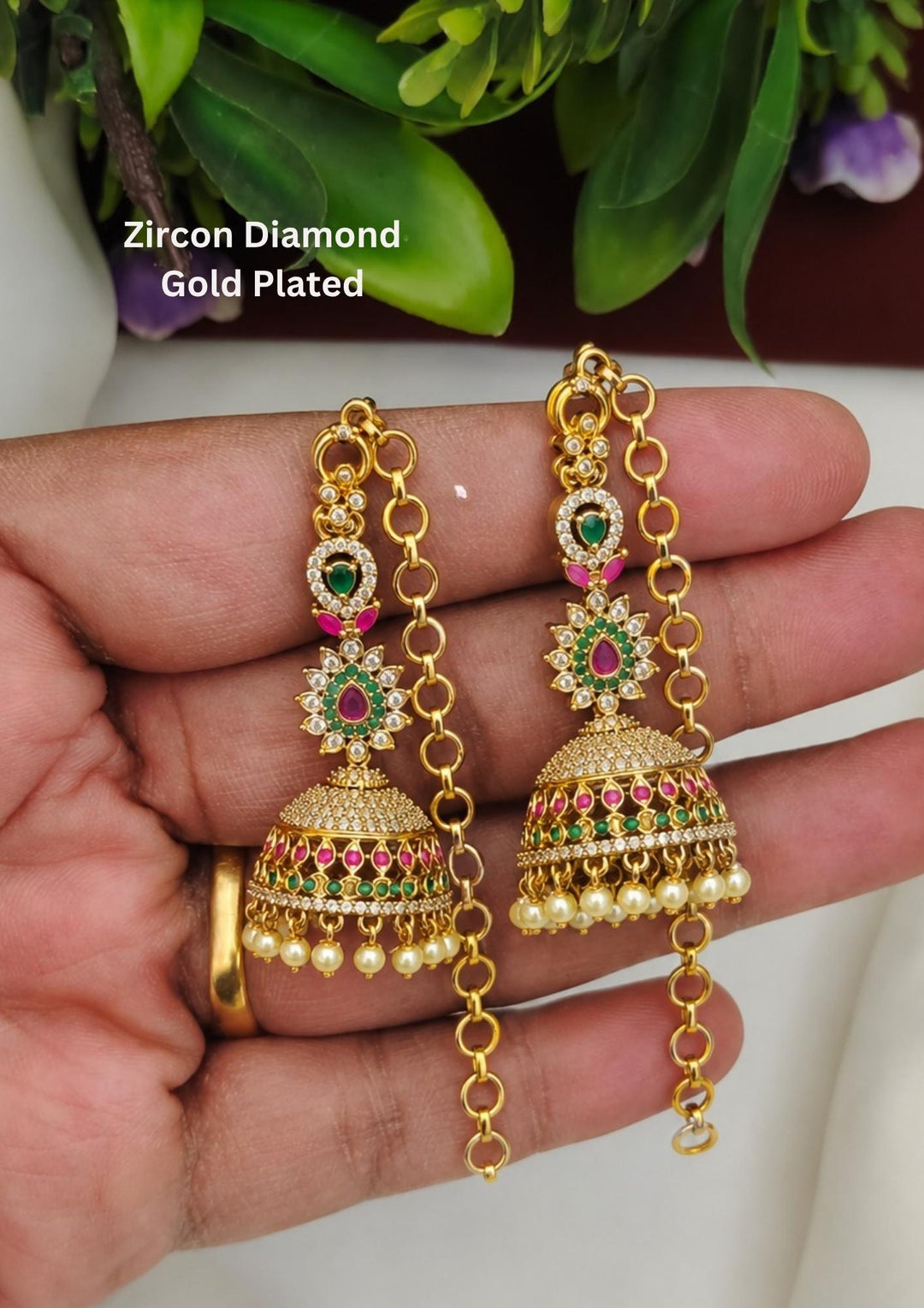 A pair of traditional gold plated jhumka earrings featuring bell-shaped drops tightly paved with glittering micro zircon crystals. The floral studs and dome trim are inlaid with tiny ruby pink and emerald green stones, while the bottom rim is finished with delicate hanging cream pearls. Attached are matching classic golden link hair chains.