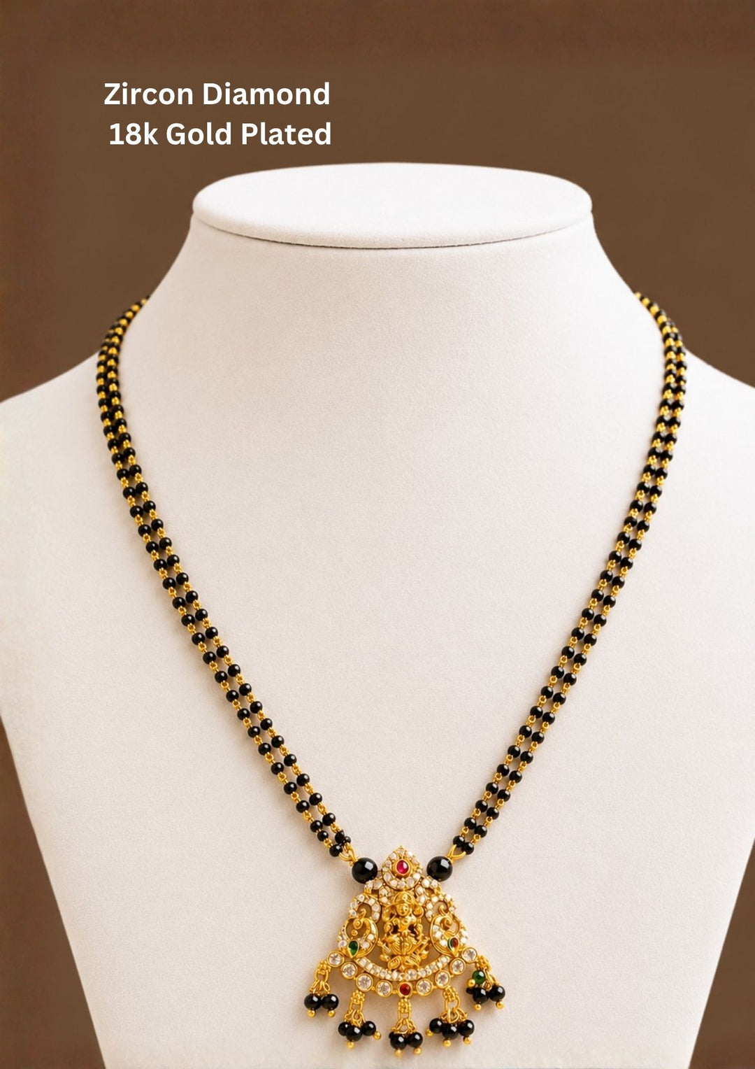 A traditional 18k gold plated mangalsutra necklace featuring a double strand of classic small black beads. The centerpiece is a half-moon shaped temple pendant intricately embossed with a seated Goddess Lakshmi motif framed by peacocks, accented with sparkling white zircon crystals, ruby-red drops, and hanging black bead clusters, perfect for weddings, bridal wear, functions. it is anti fade color and skin friendly