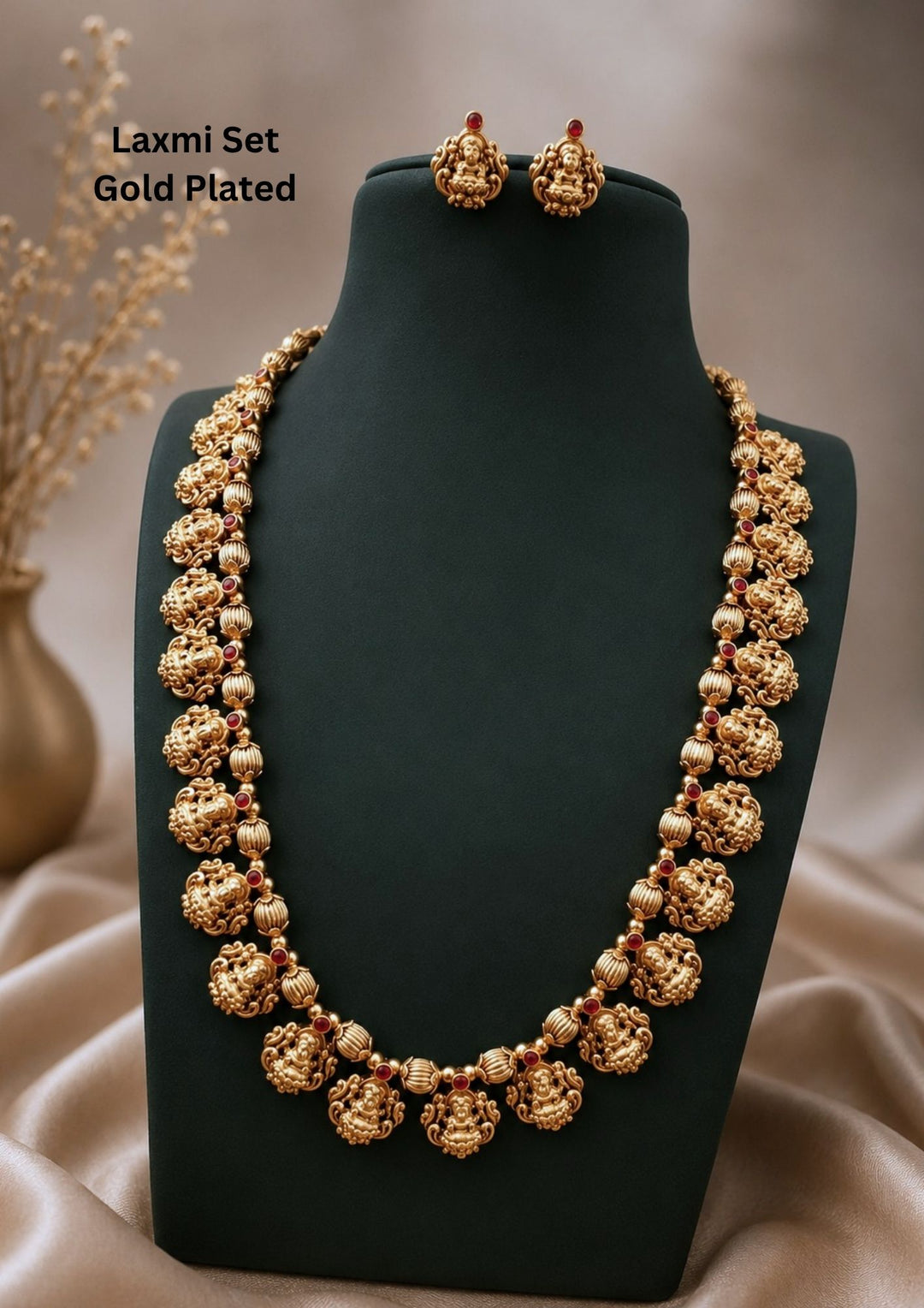 gold platedbrass base temple jewelry haram set featuring a long traditional chain structured with a continuous row of beautifully carved Goddess Lakshmi motifs seated on lotus bases, interspersed with ribbed gold beads and tiny pink ruby stone spacers. The set includes matching small temple stud earrings featuring the same seated Lakshmi design with a top ruby accent. Perfect for weddings, festivals, and traditional family functions with skin-friendly premium quality, comfortable fitting