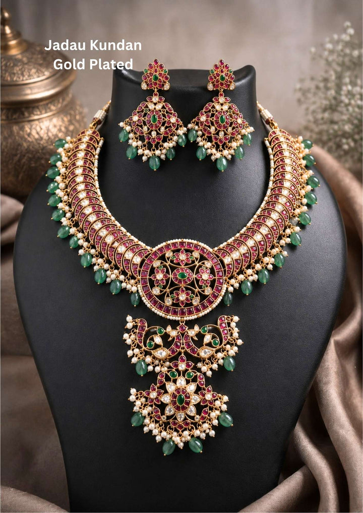 Gold plated Jadau Kundan jewelry set in image_e6bd29.png featuring a broad, curved collar choker necklace meticulously detailed with red kemp-style fills and tiny pearl borders. The center showcases a large circular medallion connected to a multi-tiered dangling floral pendant. The entire lower fringe of the necklace is accented with soft green bead tumbles and delicate pearl clusters. Includes a matching pair of heavy, diamond-shaped hanging earrings. Perfect for weddings and festivals