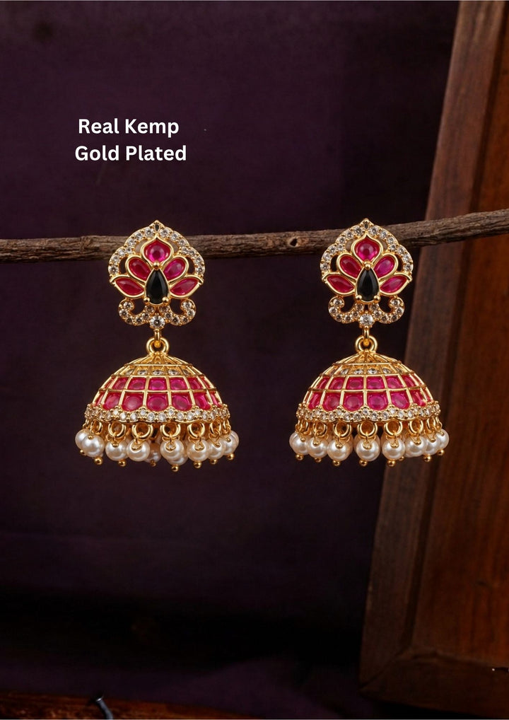 Gold plated jhumka earrings featuring an ornate, peacock-inspired floral top stud set with a central black teardrop stone, vibrant ruby-pink petals, and a border of sparkling white CZ diamonds. The hanging dome-shaped jhumka bell is meticulously lined with rows of ruby-pink stones and white crystals, finished with a delicate fringe of dangling white pearls. Crafted for festive celebrations with premium quality