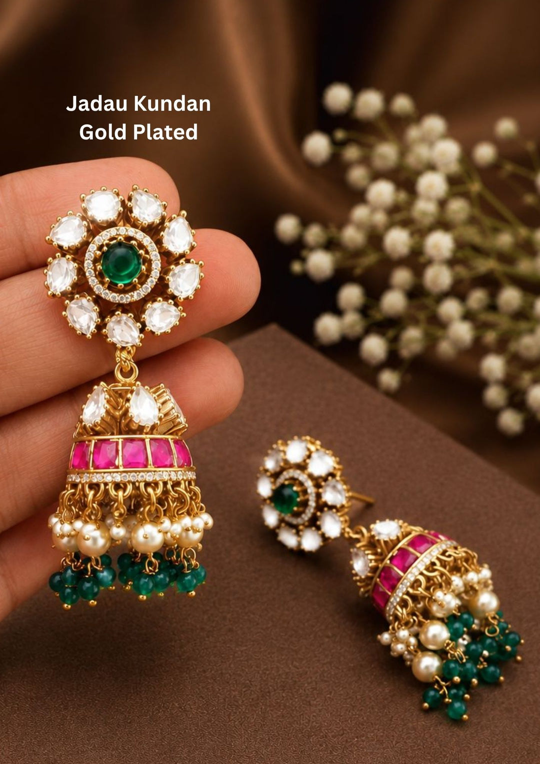A pair of heritage gold plated jhumka earrings featuring a large round floral top stud filled with uncut white kundan stones and a deep green center stone. The hanging bell-shaped dome is decorated with a hot pink enamel band, white pearl fringes, and dangling dark green emerald-color beads. These statement earrings are built with premium materials and secure post backings.