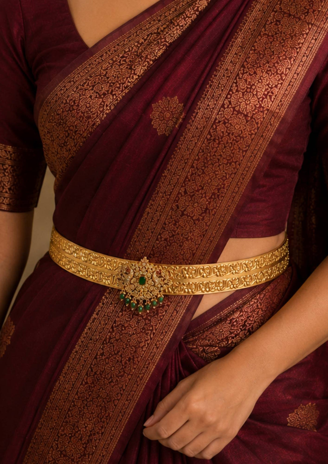 A close-up of a model wearing a Elegant gold plated traditional hipbelt featuring floral engraved patterns with a grand peacock-inspired centerpiece embellished with sparkling zircon stones, an emerald green center stone and green bead danglers.
