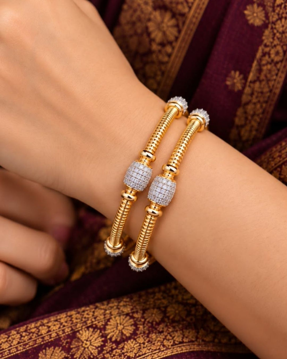 Pair of gold plated designer bangles displayed on a dark wooden surface against a black fabric background.