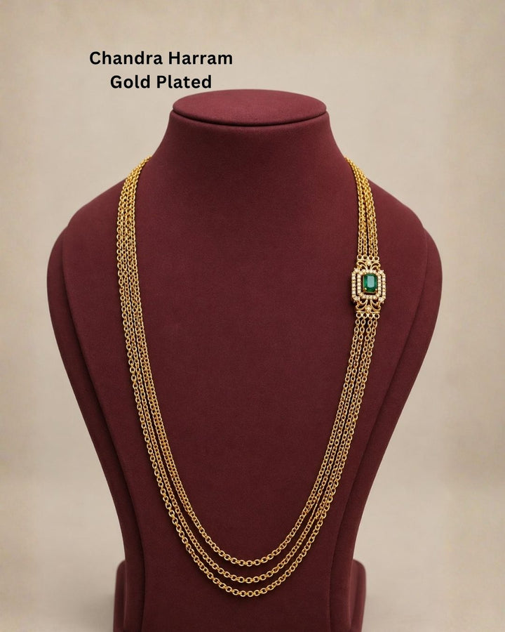 Beautiful green stone Chandra Haram gold plated necklace featuring multiple layered chains and an elegant rectangular green centerpiece with intricate detailing and sparkling stones. Perfect for festive occasions, traditional outfits, weddings, temple visits, and premium gifting.