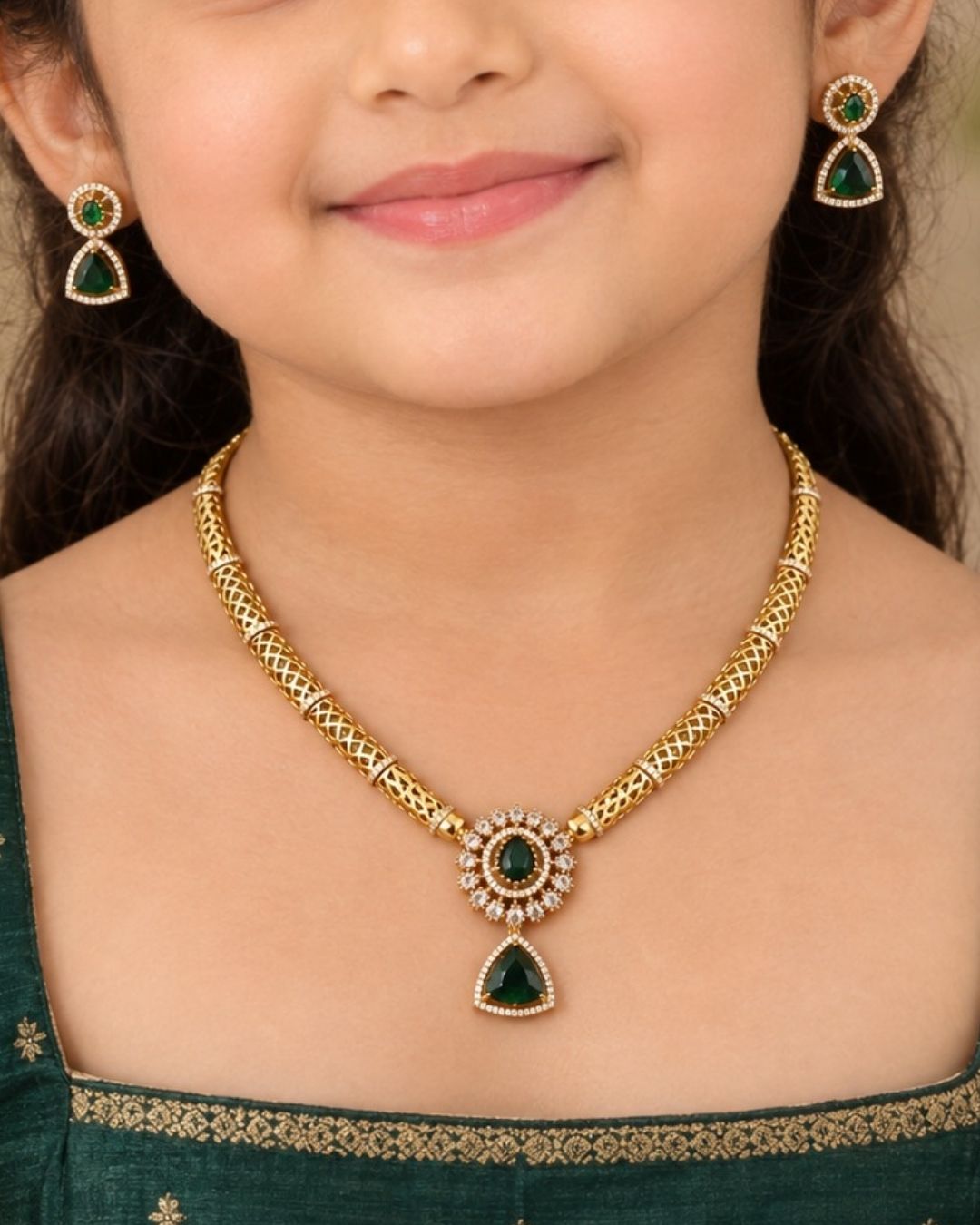 Gold-plated necklace and matching earrings set displayed on a black jewelry bust. The necklace features a textured gold chain with a central teardrop pendant and matching drop earrings, embellished with green stones and small clear stones.