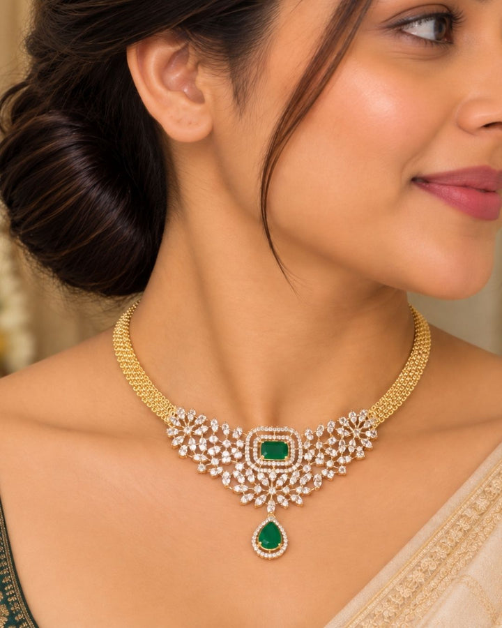 It centers an intricate, V-shaped floral bib pattern encrusted with glittering white American Diamonds (CZ stones), a prominent square emerald-green center stone, and a dangling teardrop emerald charm at the base. Includes a matching pair of elegant hanging earrings featuring a floral design with square and teardrop green stone accents. Perfect for parties and festive occasions with skin-friendly premium quality, comfortable fit,