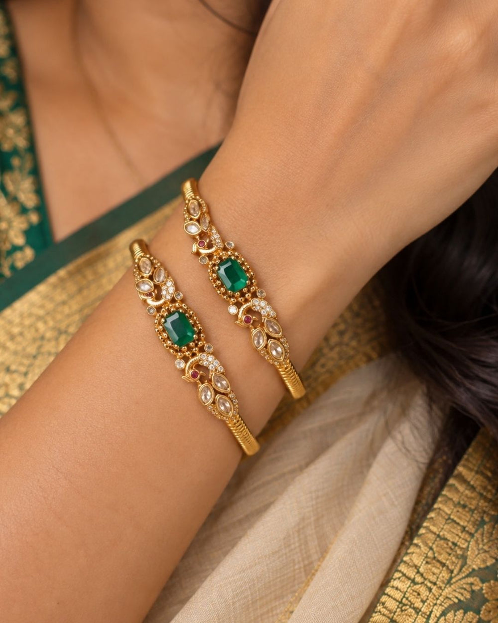 A pair of premium 18k gold plated kada bangles featuring an exquisite twin peacock design flanking a central rectangular emerald green gemstone. The peacock silhouettes are delicately studded with tiny shimmering white stones and accented with ruby red eyes. The remaining portion of the band features a clean, highly polished ribbed texture.