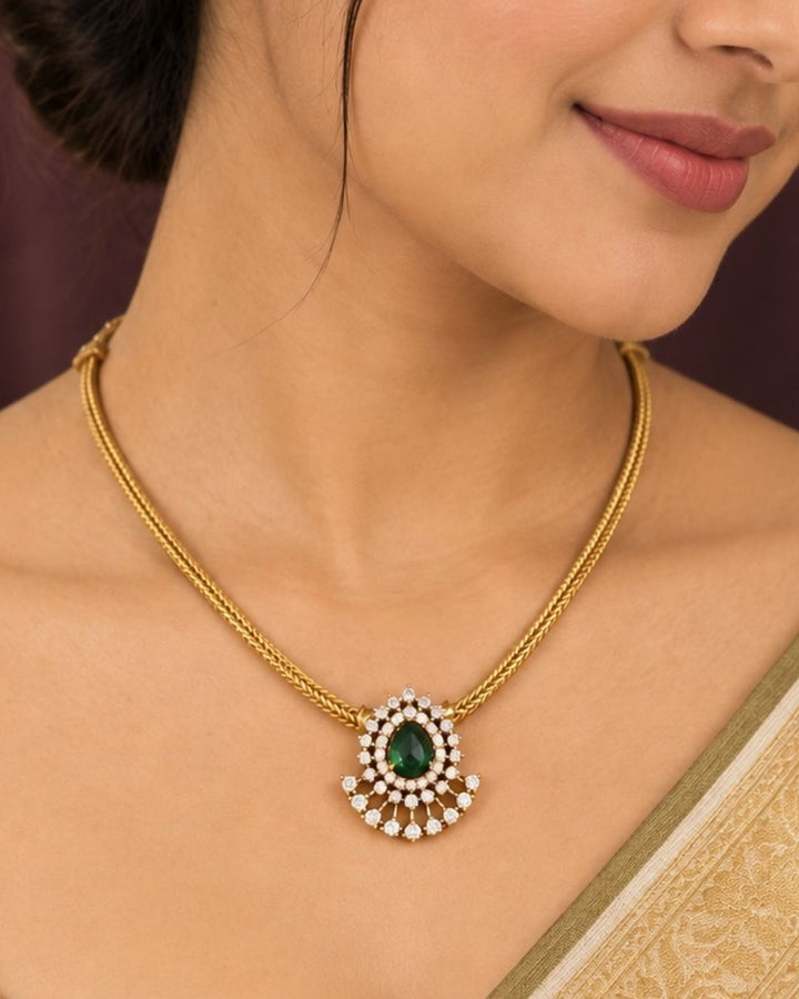 A minimalist 18k gold plated herringbone chain necklace featuring a central teardrop-shaped ruby green stone. The stone is enclosed in a halo of white pure Zircon diamonds that flares out at the bottom into a shimmering, fan-shaped scalloped fringe, paired with matching crown-set ear studs.