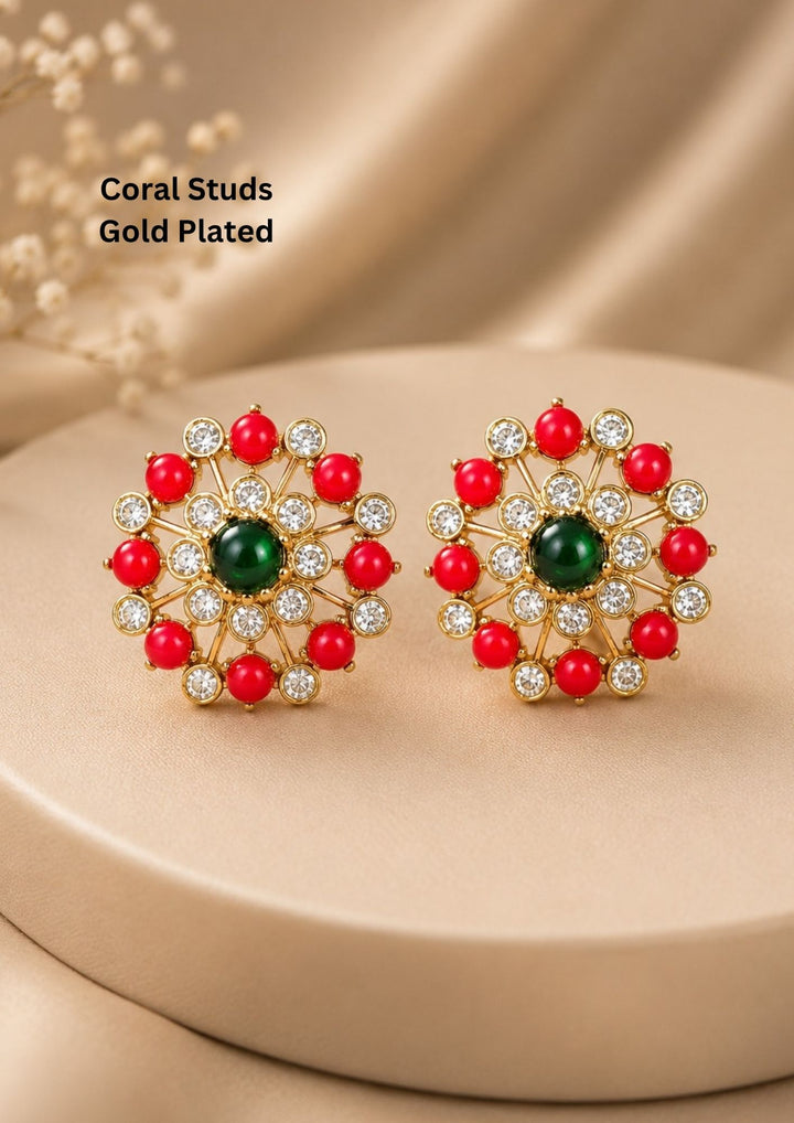 A pair of gold-plated circular stud earrings featuring a vibrant, smooth green cabochon stone at the center. The center stone is encircled by a delicate inner ring of clear cubic zirconia crystals, leading outward via gold spokes to a striking outer perimeter. This outer edge alternates between polished clear crystals and smooth, round red coral beads, creating a beautiful floral wheel design. suitable for weddings, poojas and bridal occasions.