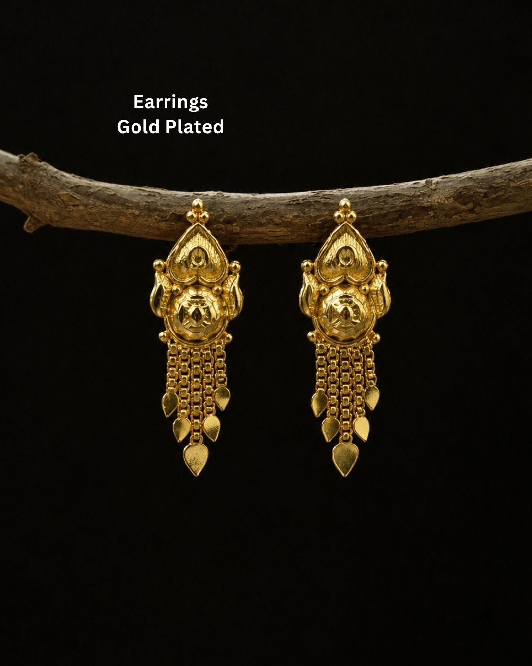 A pair of gold-plated traditional Indian jhumka earrings hanging from a rustic wooden branch against a solid black background, with white text overlay reading &