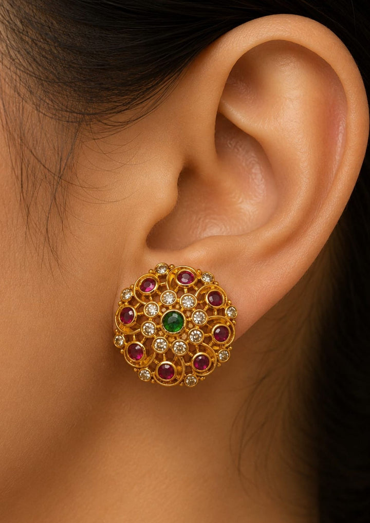 Close-up of an ear wearing a Traditional jadau kundan gold plated stud earrings featuring an intricate circular floral design crafted with radiant ruby-colored stones, sparkling kundan stones and a striking green center stone. The symmetrical pattern and antique-inspired craftsmanship create a rich ethnic appeal suitable for weddings, festivals, poojas and traditional celebrations.