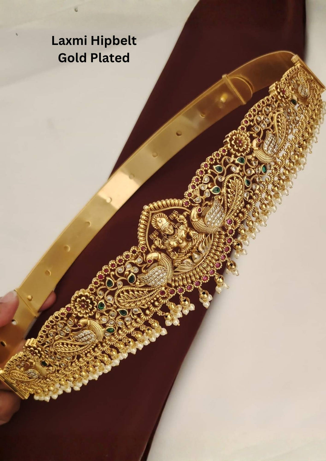 An elegant gold plated hipbelt (vaddanam) from featuring a detailed central Goddess Laxmi motif seated on a lotus. The waist belt showcases intricate nakshi craftsmanship with peacock designs, kemp stones, green accents, and delicate hanging pearl clusters along the bottom edge.
