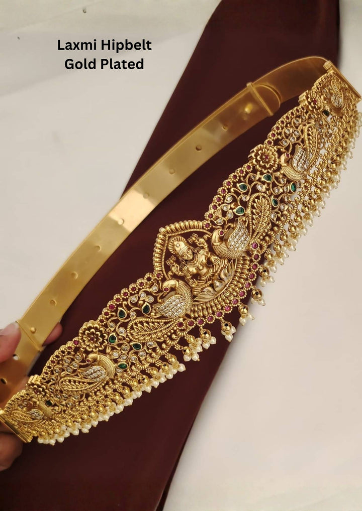 An elegant gold plated hipbelt (vaddanam) from featuring a detailed central Goddess Laxmi motif seated on a lotus. The waist belt showcases intricate nakshi craftsmanship with peacock designs, kemp stones, green accents, and delicate hanging pearl clusters along the bottom edge.
