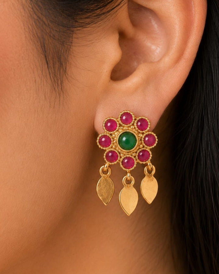 Gold-toned traditional Indian jewelry set featuring a choker necklace and matching drop earrings worn by a model. The jewelry is adorned with gold leaf-shaped charms, small beads, and central floral motifs set with green and red stones.