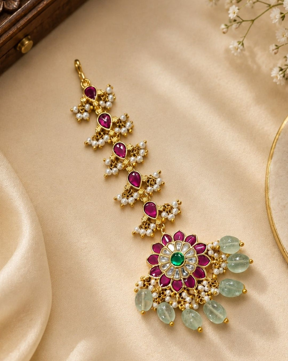 Beautiful traditional gold plated maang tikka featuring multiple pink teardrop stones along the chain, a floral pendant with vibrant pink petals and a green center stone, sparkling white stones, delicate pearl clusters, and multiple green bead drops. Perfect for weddings, festive occasions, traditional outfits, bridal styling, and special celebrations.