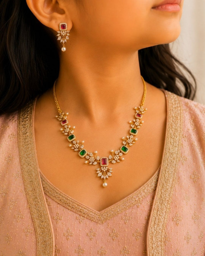 Gold-plated Navratna jewelry set featuring a necklace and matching earrings The set is adorned with gold-plated floral motifs, green rectangular stones, pink rectangular stones, and white pearl-like drops. Elegant Navaratna gold plated necklace set featuring colorful gemstone-inspired stones, pearl drops, and matching earrings for festive occasions.