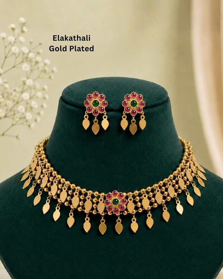 Gold-toned traditional Indian jewelry set featuring a choker necklace and matching drop earrings worn by a model. The jewelry is adorned with gold leaf-shaped charms, small beads, and central floral motifs set with green and red stones.