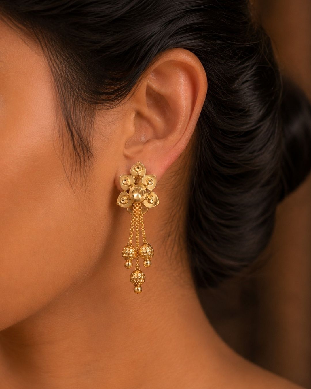 A pair of gold-plated traditional Indian jhumka earrings hanging from a wooden branch against a dark, blurred background. White text in the upper left reads &