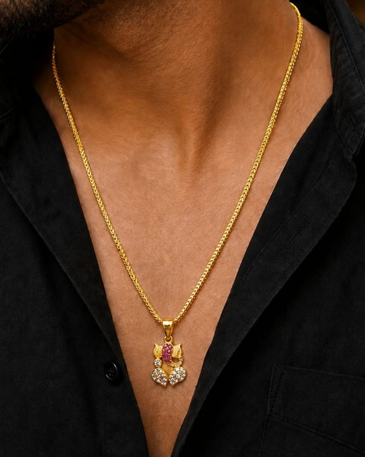 A gold-plated Ganesha pendant decorated with pink gemstones and clear stones, displayed on a dark brown carved wooden leaf with &