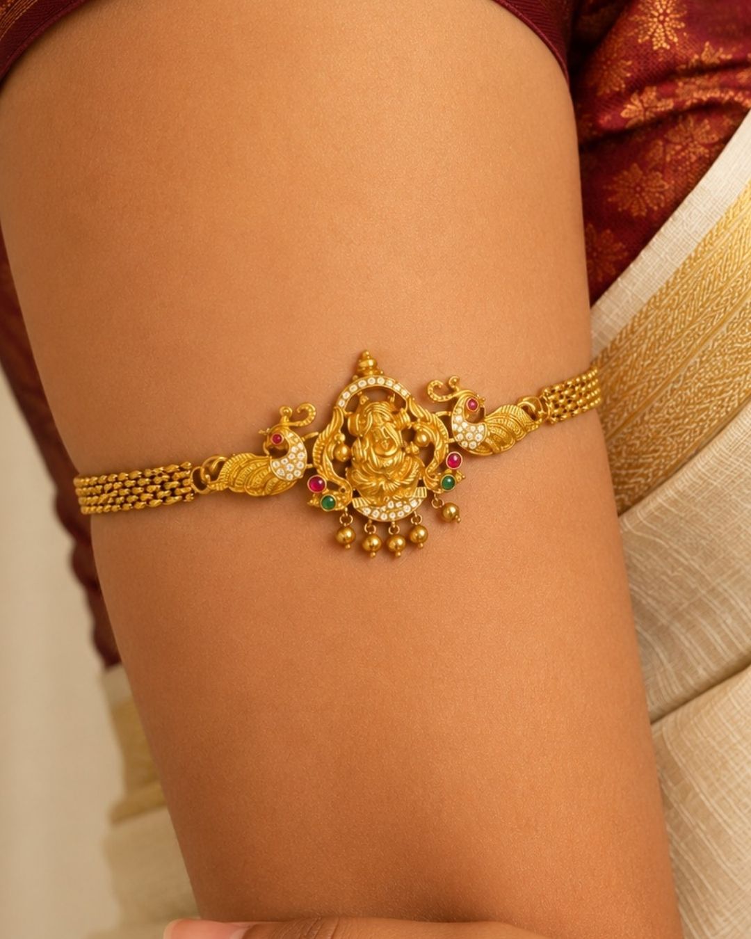 Premium gold plated choker and bajuband featuring an intricately crafted Goddess Lakshmi centerpiece surrounded by peacock motifs, white stones, red and green stone embellishments and delicate hanging gold beads. Perfect for weddings, festive occasions, traditional wear, dance performances, parties and gifting.