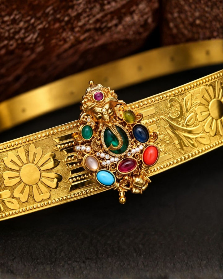 Beautiful gold plated kids hip belt featuring an intricately engraved wide band with floral designs and a central Ganesha motif surrounded by colorful green, red, blue, and white stones. Perfect for girls&
