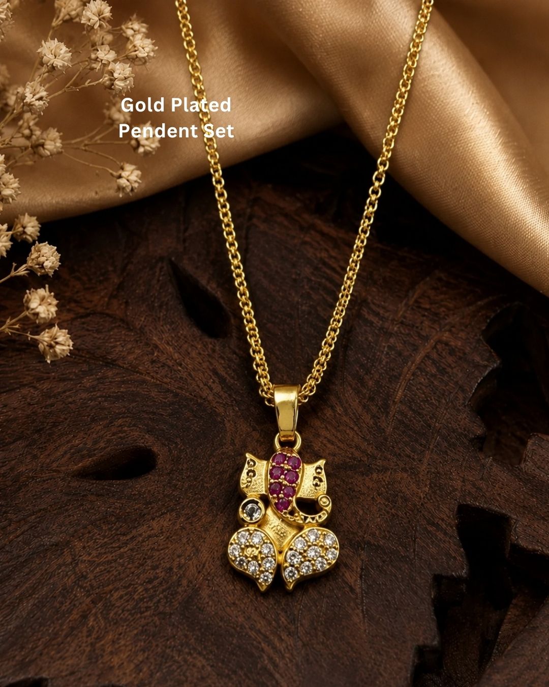 A gold-plated Ganesha pendant decorated with pink gemstones and clear stones, displayed on a dark brown carved wooden leaf with &