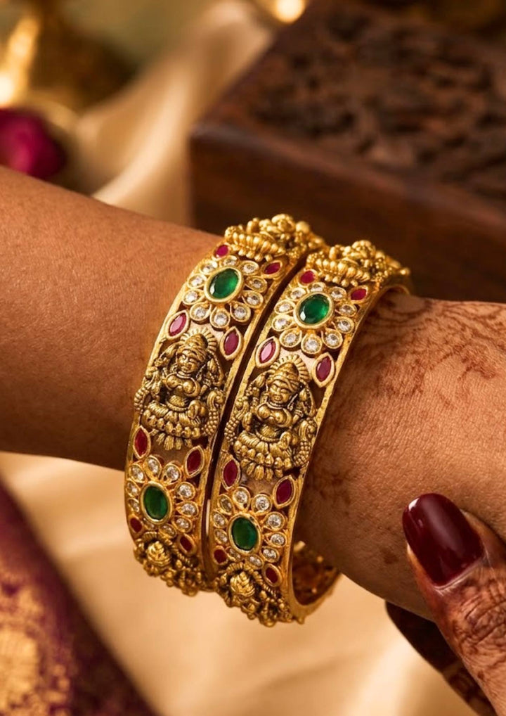 Traditional South Indian bridal attire featuring a pair of antique gold-plated Laxmi temple bangles. The jewelry showcases classic temple motifs, rich green and red stones, and white gemstone work that perfectly complements silk sarees, traditional lehengas, and bridal outfits for weddings, festivals, and special cultural occasions.