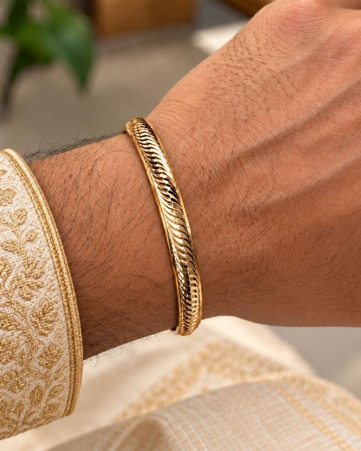 Gold plated men’s kada displayed upright on a beige surface with a soft pink background, highlighting its woven wave design and polished finish.