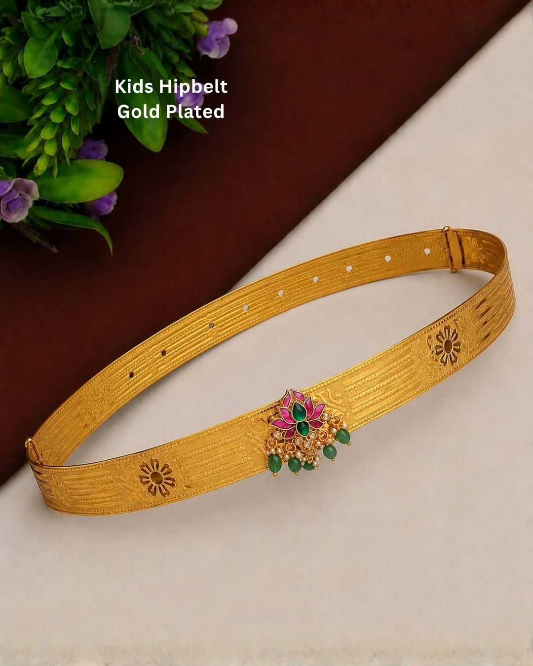 Beautiful gold plated kids hip belt featuring a wide intricately engraved band with traditional floral motifs and a decorative centerpiece adorned with vibrant green and pink stones, sparkling clear stones, and elegant green bead drops. Perfect for traditional wear, weddings, festivals, classical dance, special occasions, and gifting.