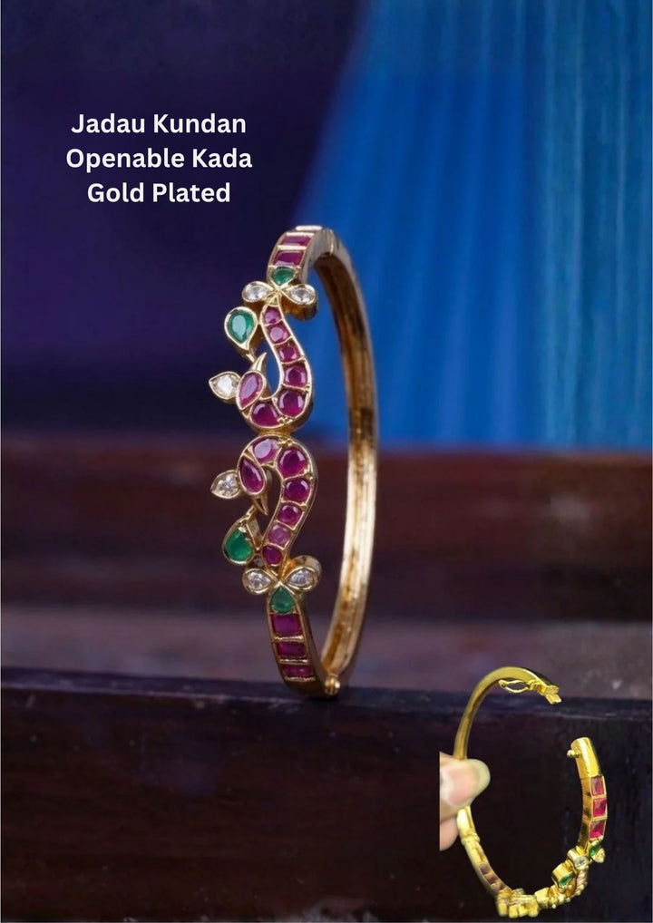 A slender 18k gold plated openable kada bangle showcasing a stylized abstract twin peacock motif along the top framework. The artistic design is inlaid with smooth cabochon ruby pink and teardrop emerald green gemstones, highlighted by clear white zircon diamond crystals at the tips and tails.