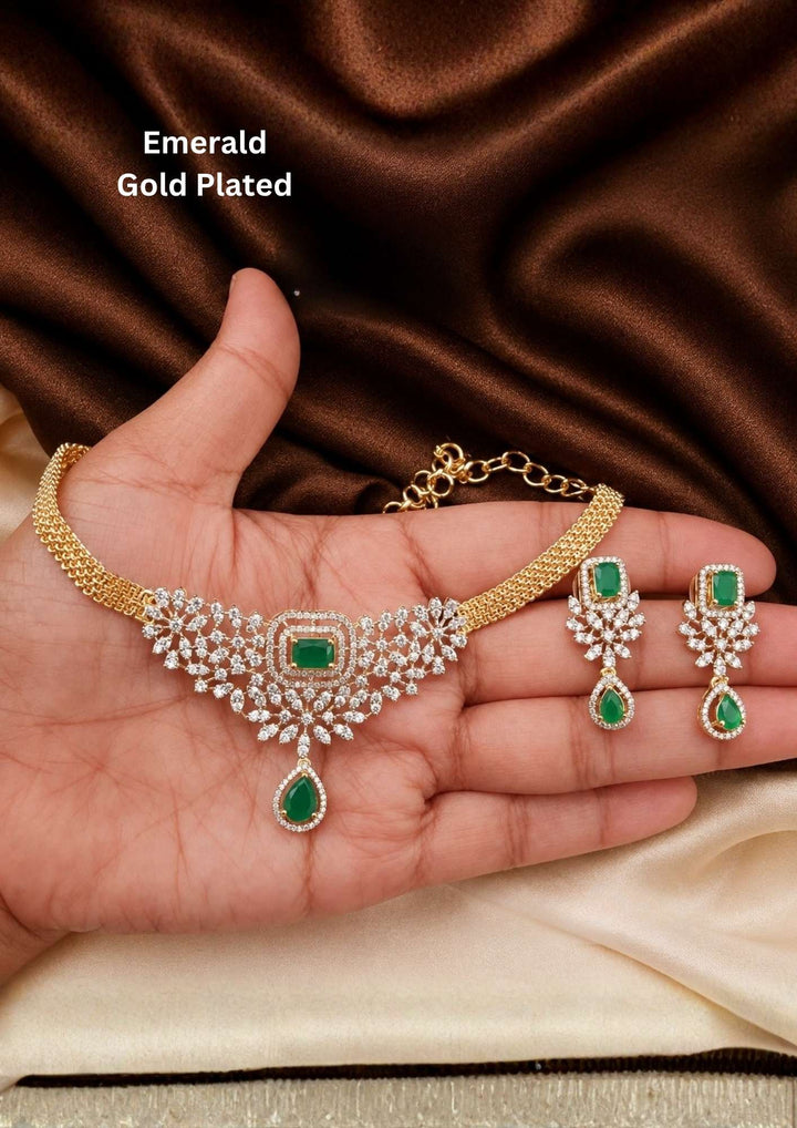 It centers an intricate, V-shaped floral bib pattern encrusted with glittering white American Diamonds (CZ stones), a prominent square emerald-green center stone, and a dangling teardrop emerald charm at the base. Includes a matching pair of elegant hanging earrings featuring a floral design with square and teardrop green stone accents. Perfect for parties and festive occasions with skin-friendly premium quality, comfortable fit,