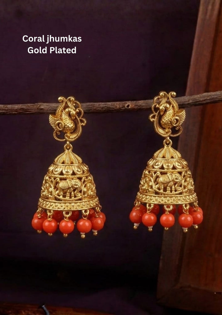 Traditional coral jhumkas gold plated earrings featuring intricate temple-inspired craftsmanship with beautifully carved elephant motifs on the dome-shaped jhumka. The earrings are adorned with vibrant coral red bead danglers and detailed antique gold finish patterns, creating a rich South Indian ethnic look suitable for weddings, festivals, poojas and cultural celebrations.
