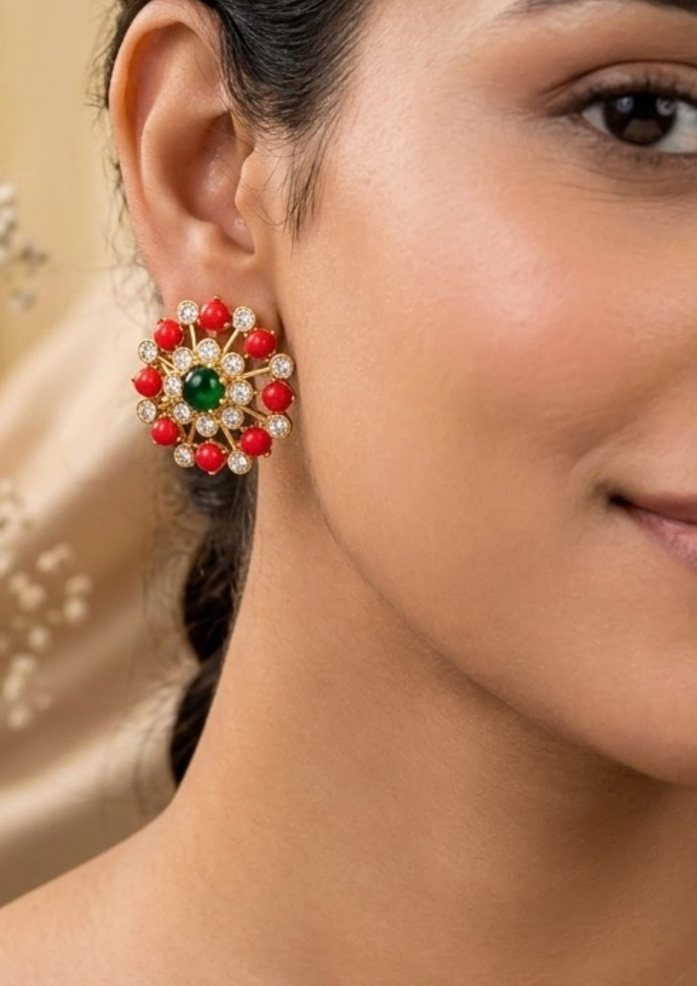 Close-up of a woman wearing colorful earrings a pair of gold-plated circular stud earrings featuring a vibrant, smooth green cabochon stone at the center. The center stone is encircled by a delicate inner ring of clear cubic zirconia crystals, leading outward via gold spokes to a striking outer perimeter. This outer edge alternates between polished clear crystals and smooth, round red coral beads, creating a beautiful floral wheel design. suitable for weddings, poojas and bridal occasions.