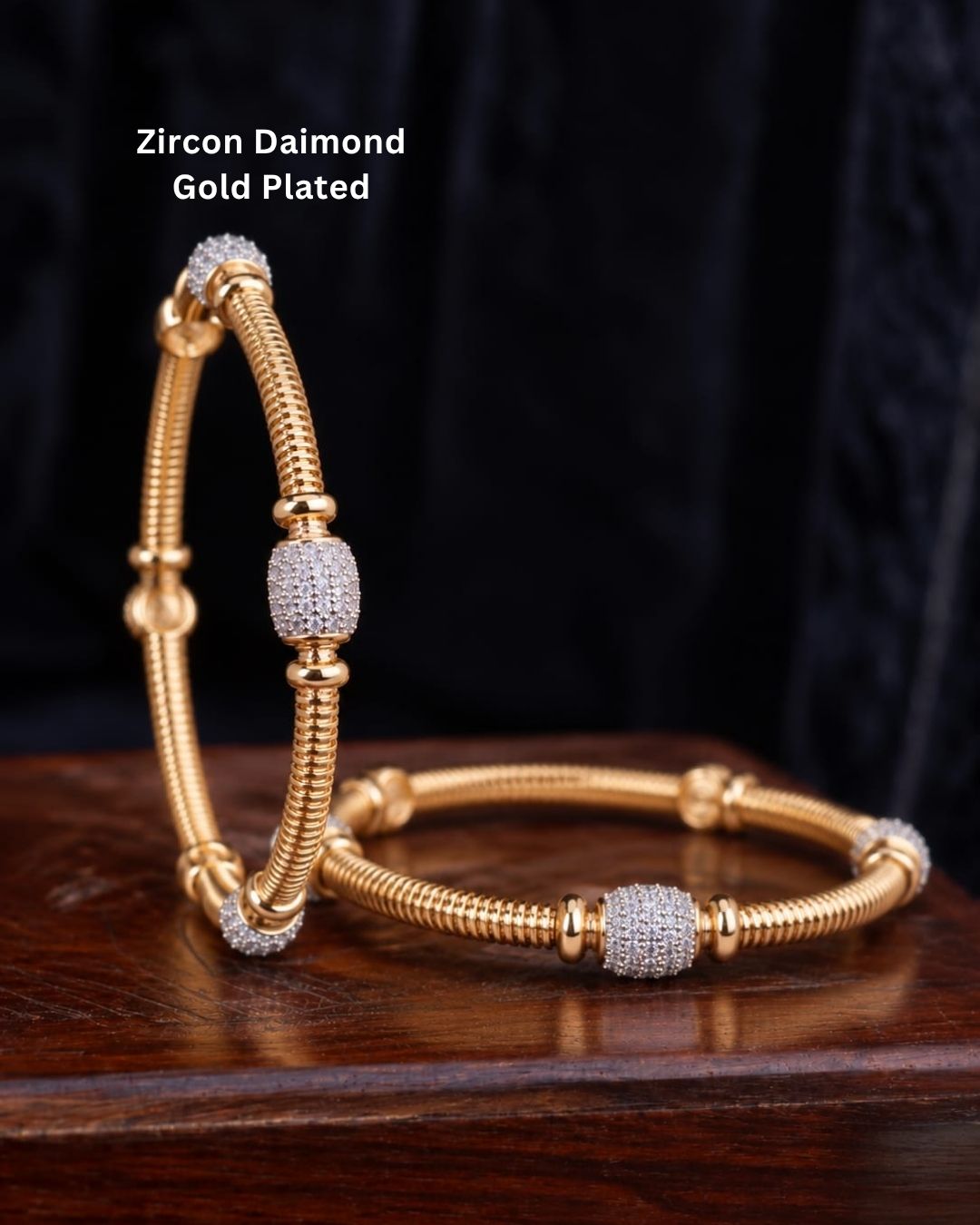 Pair of gold plated designer bangles displayed on a dark wooden surface against a black fabric background.