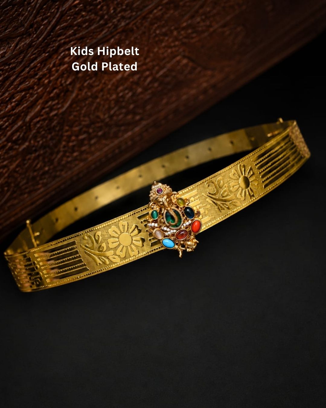Beautiful gold plated kids hip belt featuring an intricately engraved wide band with floral designs and a central Ganesha motif surrounded by colorful green, red, blue, and white stones. Perfect for girls&