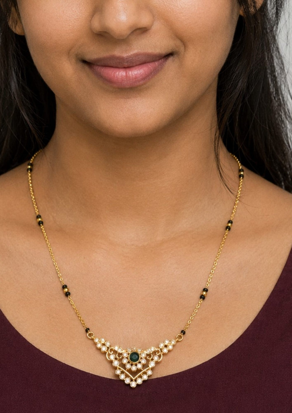 Woman wearing a gold necklace with a green gemstone Modern Zircon Diamond gold plated mangalsutra necklace The design features a delicate gold link chain transitioning into sections of traditional small black beads on both sides. The central pendant showcases a sleek, contemporary curved V-shape and a stylized butterfly or floral motif encrusted with sparkling white cubic zirconia stones. This elegant neckpiece perfect for daily wear, office wear, and casual outfits.