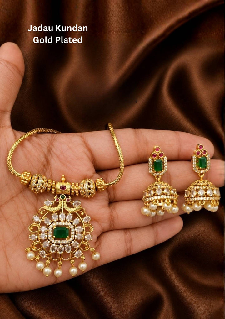 gold plated Jadau Kundan jewelry set with a sleek gold chain attached to structured gold beads. It centers a grand round medallion pendant encrusted with glittering white kundan stones, with emerald-green center stone, dual gold peacock motifs at the top, and white pearl drops at the bottom. Includes a matching pair of elegant hanging jhumka earrings Perfect for festive occasions with skin-friendly premium quality