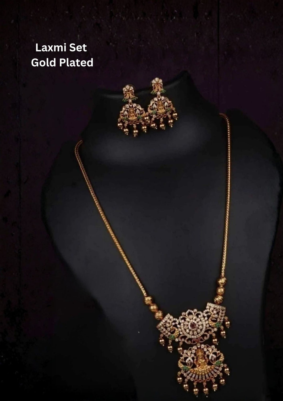 gold plated temple haram necklace set showcasing an intricate Goddess Lakshmi motif pendant flanked by detailed twin peacocks. The pendant is bordered with hanging gold beads, green emerald droplets, and white crystals, suspended from a thick rope chain and paired with matching multi-tier Lakshmi stud earrings.