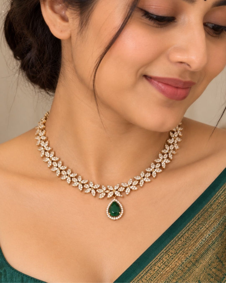 Designer gold plated necklace and earrings set featuring a delicate arrangement of leaf-shaped motifs embellished with sparkling white stones, a prominent green teardrop pendant, and matching green stone drop earrings, perfect for festive, wedding, and elegant occasions.